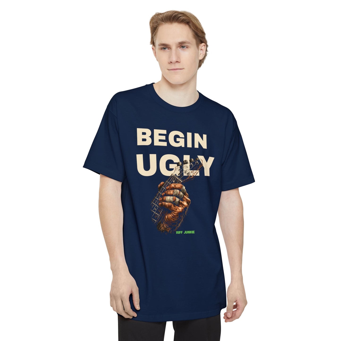 A man wearing a navy short-sleeve t-shirt featuring a graphic print on the front. The design displays the text 'BEGIN UGLY' in a distressed cream-colored font above an illustration of a hand holding a guitar, with  "Riff Junkie" in neon green.