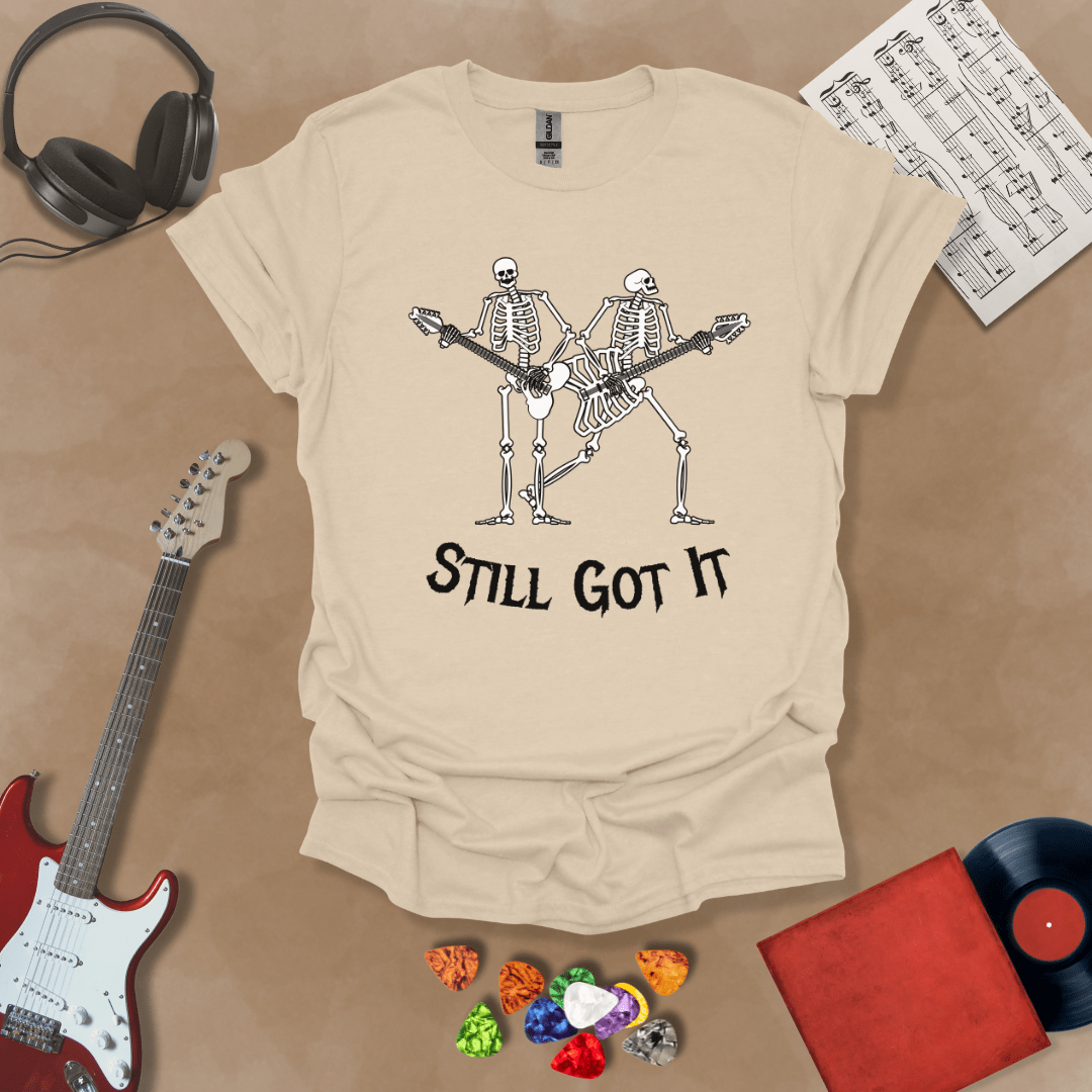 Beige Vintage style t-shirt with Two skeletons playing guitars, text 'Still Got It' with bold retro lettering and rock vibe on a transparent background.