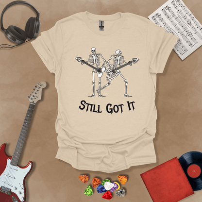 Beige Vintage style t-shirt with Two skeletons playing guitars, text 'Still Got It' with bold retro lettering and rock vibe on a transparent background.