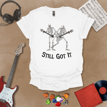 White Vintage style t-shirt with Two skeletons playing guitars, text 'Still Got It' with bold retro lettering and rock vibe on a transparent background.