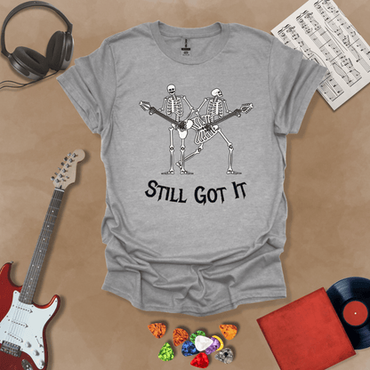 Gray Vintage style t-shirt with Two skeletons playing guitars, text 'Still Got It' with bold retro lettering and rock vibe on a transparent background.