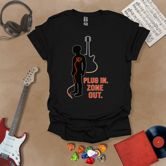 Black t-shirt with black silhouette figure, orange guitar cable plugged into heart, guitar outline behind, text “Plug In. Zone Out.”
