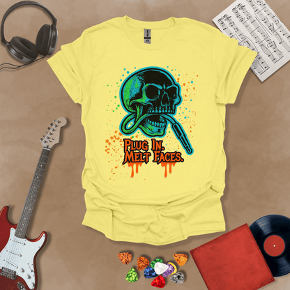 Yellow t-shirt with neon-colored skull biting a guitar cable, paint splatter effect, text “Plug In. Melt Faces.”