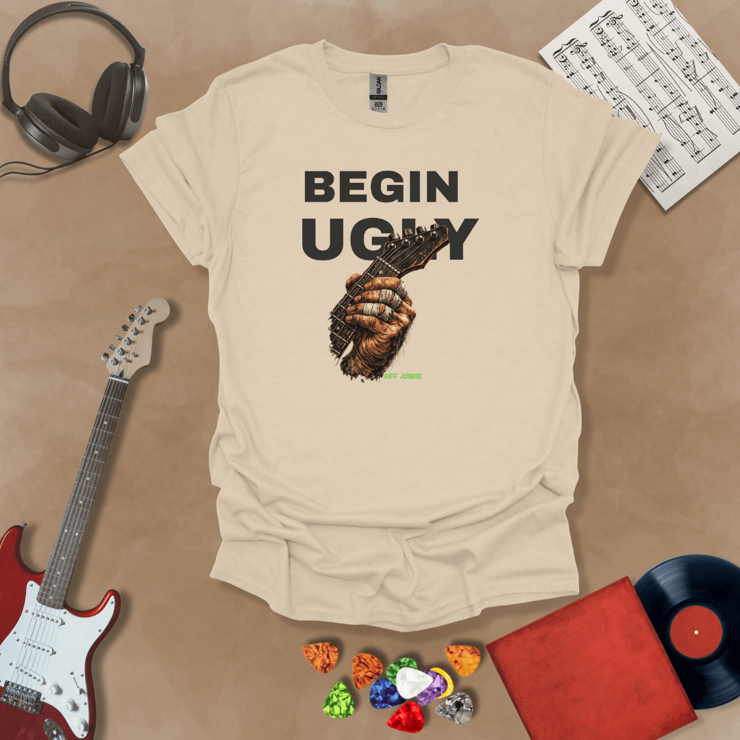 Begin Ugly – Riff Edition beige graphic t-shirt featuring bold stacked text above a weathered hand gripping a guitar fretboard with taped fingers and subtle Riff Junkie branding.