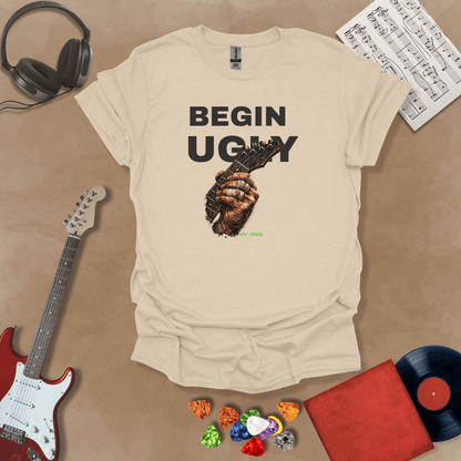 Begin Ugly – Riff Edition beige graphic t-shirt featuring bold stacked text above a weathered hand gripping a guitar fretboard with taped fingers and subtle Riff Junkie branding.