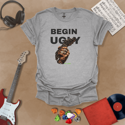Begin Ugly – Riff Edition gray graphic t-shirt featuring bold stacked text above a weathered hand gripping a guitar fretboard with taped fingers and subtle Riff Junkie branding.