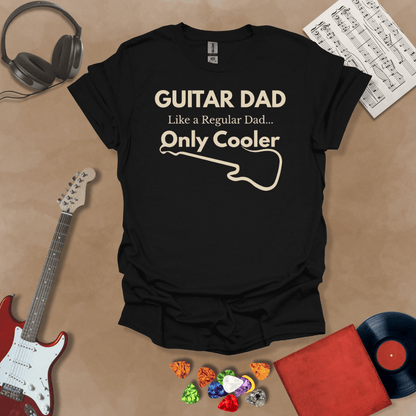A black Guitar Dad Signature Edition t-shirt laid flat on a tan background, surrounded by guitar-themed props. The design reads "GUITAR DAD – Like a Regular Dad, Only Cooler."