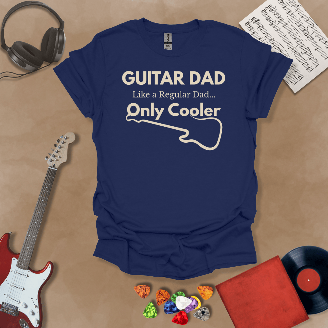 A navy Guitar Dad Signature Edition t-shirt laid flat on a tan background, surrounded by guitar-themed props. The design reads "GUITAR DAD – Like a Regular Dad, Only Cooler."