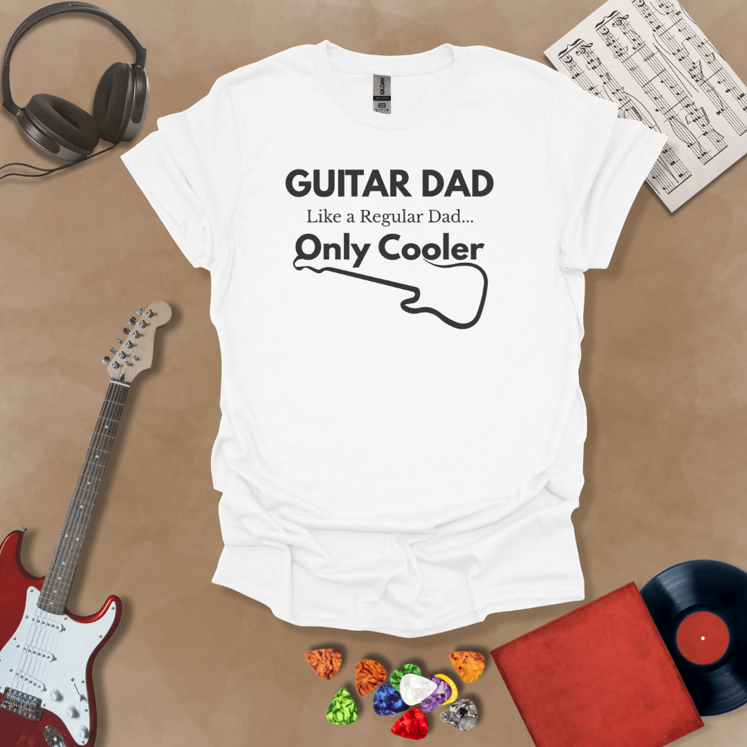 A white Guitar Dad Signature Edition t-shirt laid flat on a tan background, surrounded by guitar-themed props. The design reads "GUITAR DAD – Like a Regular Dad, Only Cooler."
