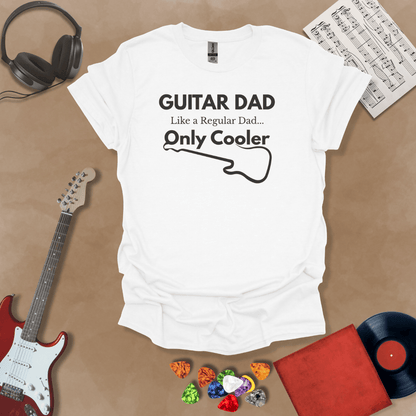 A white Guitar Dad Signature Edition t-shirt laid flat on a tan background, surrounded by guitar-themed props. The design reads "GUITAR DAD – Like a Regular Dad, Only Cooler."