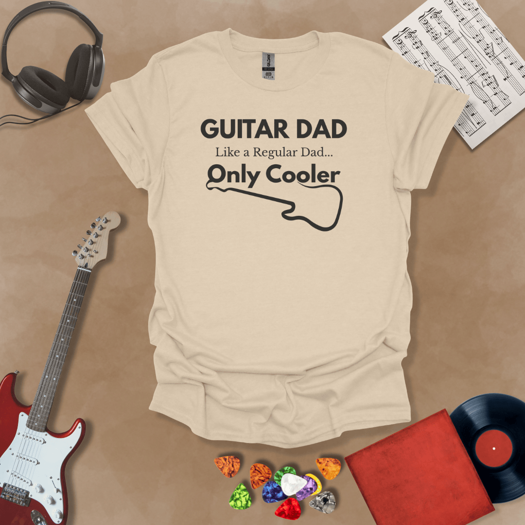 A beige Guitar Dad Signature Edition t-shirt laid flat on a tan background, surrounded by guitar-themed props. The design reads "GUITAR DAD – Like a Regular Dad, Only Cooler."