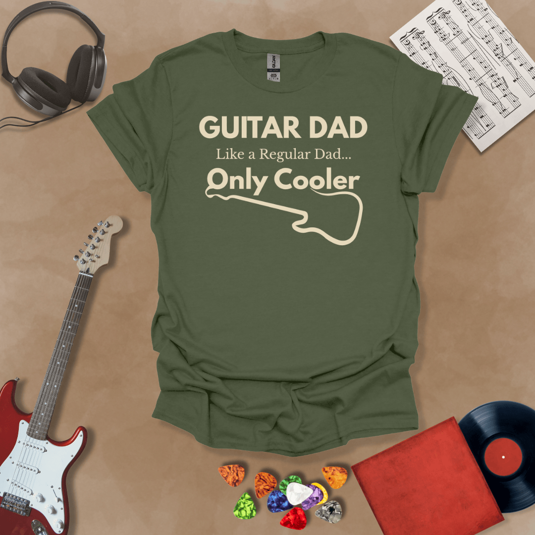 A military green Guitar Dad Signature Edition t-shirt laid flat on a tan background, surrounded by guitar-themed props. The design reads "GUITAR DAD – Like a Regular Dad, Only Cooler."