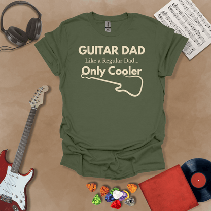 A military green Guitar Dad Signature Edition t-shirt laid flat on a tan background, surrounded by guitar-themed props. The design reads "GUITAR DAD – Like a Regular Dad, Only Cooler."