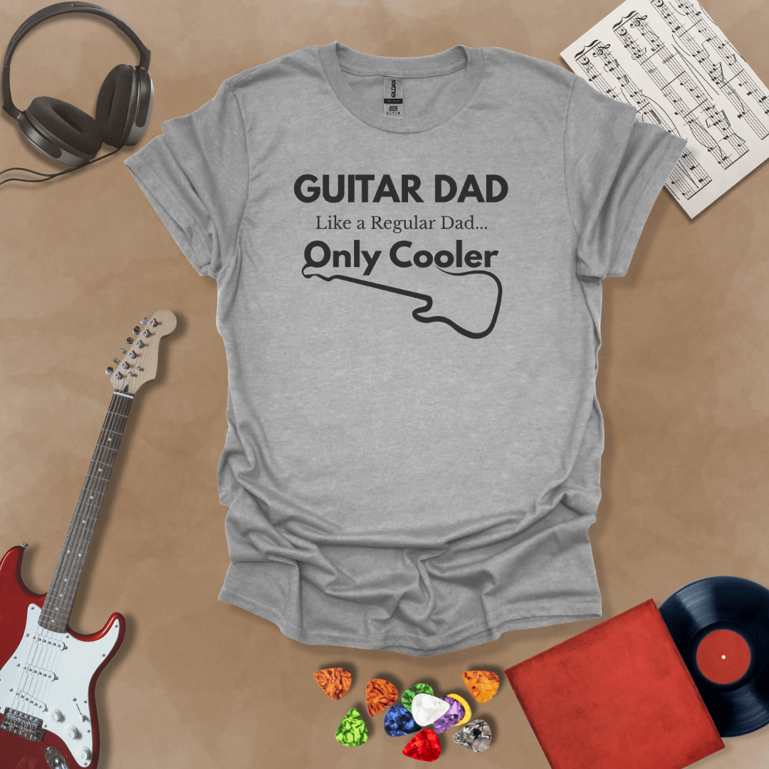 A light gray Guitar Dad Signature Edition t-shirt laid flat on a tan background, surrounded by guitar-themed props. The design reads "GUITAR DAD – Like a Regular Dad, Only Cooler."