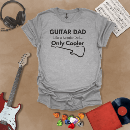 A light gray Guitar Dad Signature Edition t-shirt laid flat on a tan background, surrounded by guitar-themed props. The design reads "GUITAR DAD – Like a Regular Dad, Only Cooler."