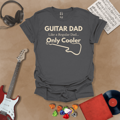A dark gray Guitar Dad Signature Edition t-shirt laid flat on a tan background, surrounded by guitar-themed props. The design reads "GUITAR DAD – Like a Regular Dad, Only Cooler."