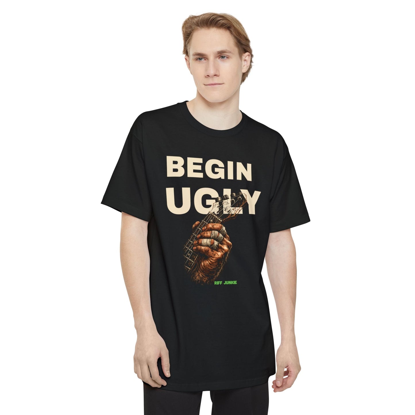 A man wearing a black short-sleeve t-shirt featuring a graphic print on the front. The design displays the text 'BEGIN UGLY' in a distressed cream-colored font above an illustration of a hand holding a guitar, with  "Riff Junkie" in neon green.